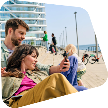 Lächelndes Paar mit Smartphone entspannt auf Strandpromenaden-Stufen, Passanten im Hintergrund.