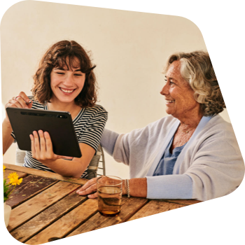 Junge Frau und ältere Dame lachen zusammen beim Betrachten eines Tablets an einem Holztisch.